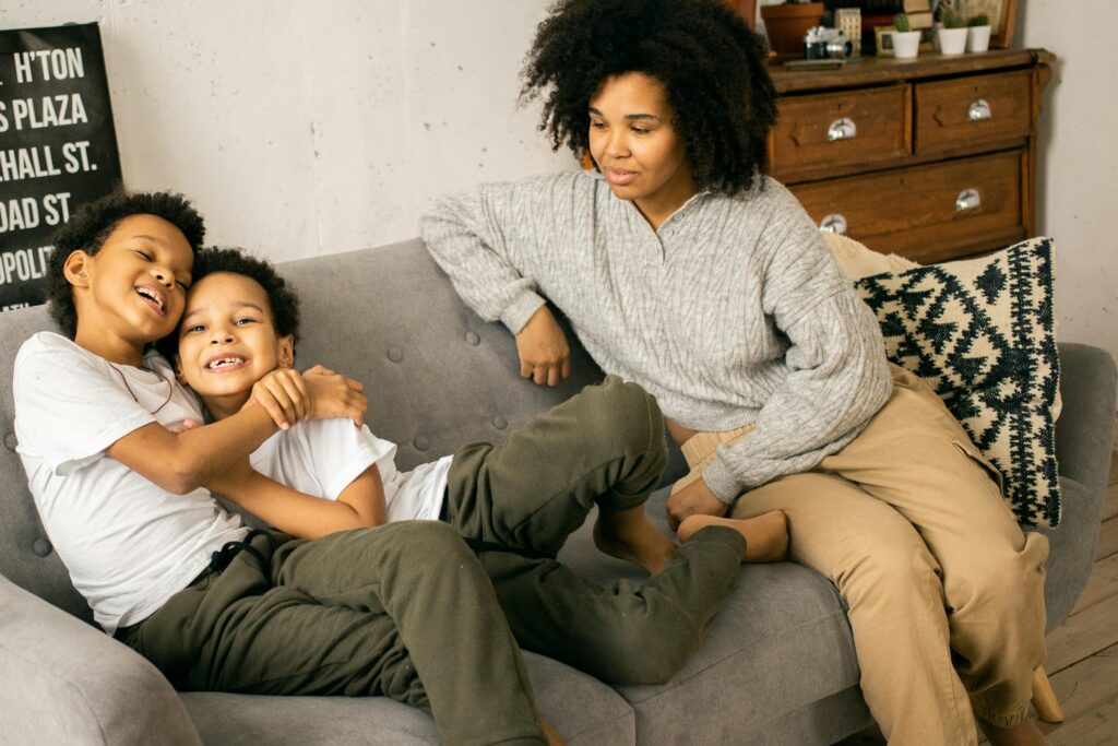 Cheerful African American boys in casual wear hugging on comfortable sofa with caring black mother in light room with wooden cupboard