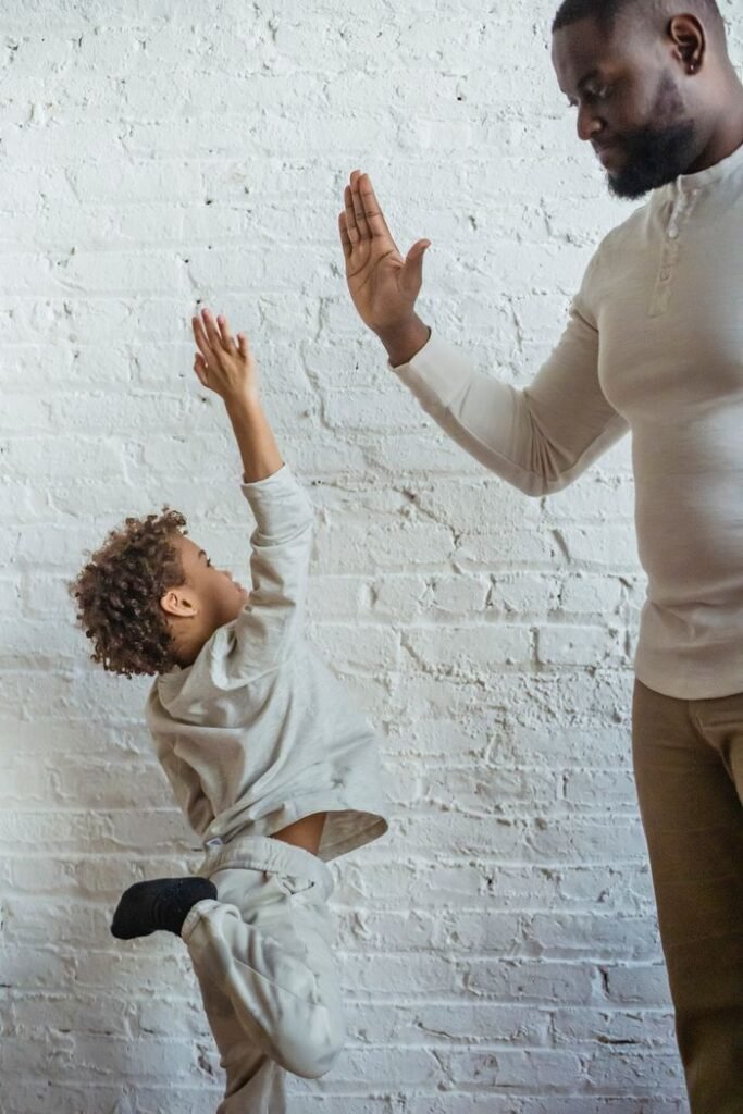 Positive adult African American father and cute little son giving high five and standing against white brick wall