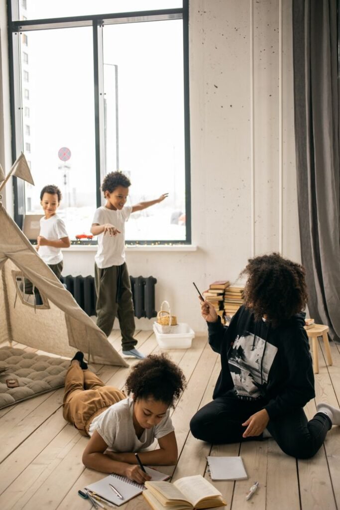 African American mother helping daughter with homework while sitting on wooden floor near sons playing in room near blanket fort