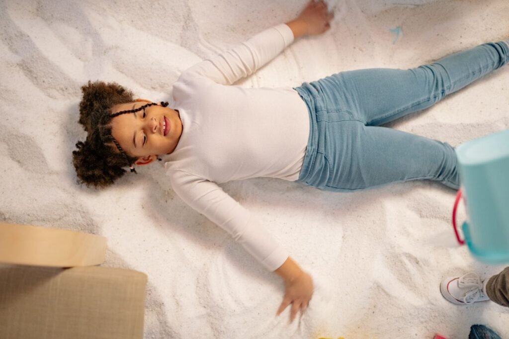Young girl lying down on sand indoors with a joyful expression, engaging in mindful play.